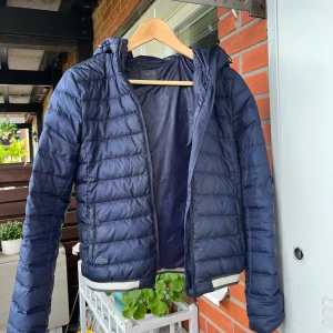 Mörkblå pufferjacka från Lacoste - Snygg mörkblå pufferjacka från Lacoste med huva och dragkedja. Jackan är quiltad och har en sportig vibe med diskret logga på bröstet. Tillverkad i polyester och perfekt för kyliga dagar. Passformen är normal och jackan har långa ärmar. En liten 36:a. Behöver kemtvättas, kolla tvättlapp. 