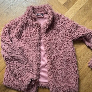Rosa teddyjacka från Gina Tricot - Fluffig teddyjacka i dammigt rosa från Gina Tricot. Jackan har lång luddig pälslook! Perfekt för dig som vill sticka ut och hålla dig varm med stil. Snygg att slänga över en hoodie eller t-shirt.