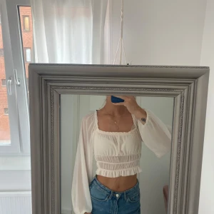 Vit croppad blus med volang - Superfin vit blus med croppad passform och långa, luftiga ärmar. Toppen har fyrkantig ringning, smockad midja och volangdetaljer både upptill och nedtill. Perfekt för dig som gillar romantisk och trendig stil.