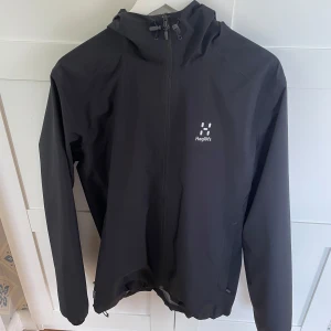 Svart vindjacka från Haglöfs med huva - Svart vindjacka från Haglöfs med minimalistisk design och justerbar huva. Jackan har dragkedja framtill, två sidofickor och diskret logga på bröstet. Perfekt för blåsiga dagar och enkel att matcha med allt. Snygg och funktionell för uteliv.