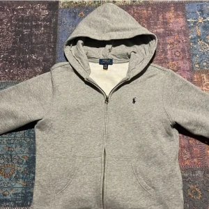 Grå hoodie från Polo Ralph Lauren - Klassisk grå hoodie från Polo Ralph Lauren med dragkedja och huva. Tröjan har broderad logga på bröstet, känguruficka och är tillverkad i mjuk bomullsmix. Perfekt för chill dagar eller när du vill ha en clean och enkel look.