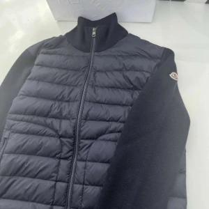 Moncler cardigan  - Snygg svart pufferjacka från Moncler med quiltad framsida och stickade ärmar. Jackan har hög krage, dragkedja framtill och Moncler-logga på ärmen. Perfekt för dig som vill ha en stilren och sportig look.