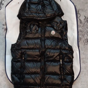 Svart dunväst från Moncler med huva - Snygg svart dunväst från Moncler med glansig finish och avtagbar huva. Västen har två dragkedjefickor framtill, quiltad design och klassisk Moncler-logga på bröstet. Perfekt för lager-på-lager och streetstyle.