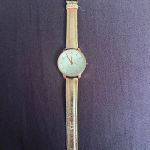 Klocka från SVR med syntetband - Stilren klocka från SVR med rund urtavla. Armbandet är i guldig syntet och har klassisk spänne. Urtavlan är enkel med tunna guldiga markeringar och visare. Perfekt accessoar för dig som gillar en clean look.