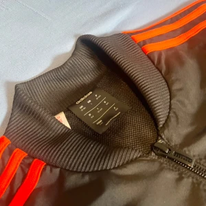 Svart och röd träningsjacka från Adidas - Snygg svart och röd träningsjacka från Adidas med de klassiska tre ränderna i rött längs ärmarna. Jackan har en dragkedja framtill och ribbade muddar vid ärmslut och krage. Perfekt för sportiga aktiviteter eller en avslappnad stil.