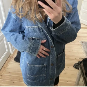 Blå jeansjacka med fickor från Monki - Säljer en cool blå jeansjacka med stora fickor framtill och knappar. Jackan har en avslappnad passform och långa ärmar. Från monki i st S och är väldigt lite använd 🥰🙌🏼☀️