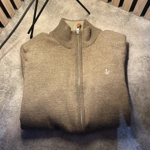 Beige stickad tröja från morris  - Säljer en stilren beige stickad tröja från morris med dragkedja och en liten broderad detalj på bröstet. Perfekt för kyliga dagar med sin höga krage och mjuka material. Passar både till vardags och mer uppklädda tillfällen.