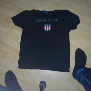 Svart t-shirt från GANT - Säljer en svart t-shirt från GANT med korta ärmar och ett tryck av GANT-loggan och en sköld med stjärnor och ränder på framsidan. Perfekt för en stilren och avslappnad look. Storlek Small är prutbar vid snabbaffär skriv om du har några frågor eller funderingar 