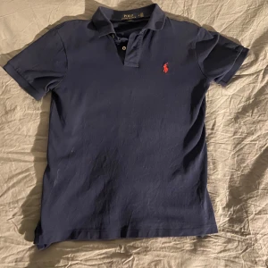 Mörkblå pikétröja från Ralph Lauren - Säljer en klassisk mörkblå pikétröja från Ralph Lauren med den ikoniska röda loggan på bröstet. Tröjan har korta ärmar och en stilren krage med knappar. Perfekt för en avslappnad men stilfull look.