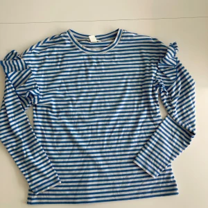 Blå- och vitrandig tröja med volangdetaljer - Säljer en blå- och vitrandig tröja med volangdetaljer på ärmarna. Tröjan har långa ärmar och en normal passform. Perfekt för en stilren och lekfull look. Hör av dig vid intresse! 💙