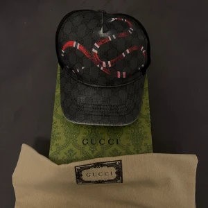Svart keps med ormmönster från Gucci - Snygg svart keps från Gucci med ett coolt ormmönster i rött och vitt. Kepsen har ett klassiskt Gucci-mönster i bakgrunden och justerbar passform. Perfekt för att ge din outfit en lyxig touch. Pris kan diskuteras