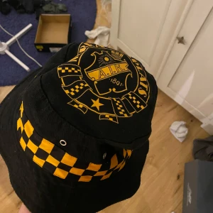 Svart och gul AIK-hatt - Snygg svart hatt med AIK-logga och gulrutigt mönster. 