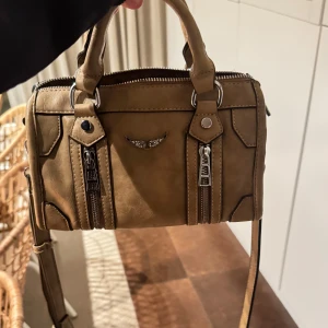 Brun axelväska med dragkedja - Snygg brun axelväska med flera dragkedjor och ett dekorativt emblem på framsidan. Väskan har både handtag och en justerbar axelrem för flexibilitet. Perfekt för att bära dina dagliga nödvändigheter med stil.