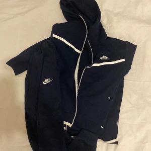 Svart träningsset från Nike - Snyggt svart träningsset från Nike med vit dragkedja och detaljer. Jackan har huva och långa ärmar, medan byxorna har elastisk midja för extra komfort. Perfekt för träning eller en avslappnad dag.