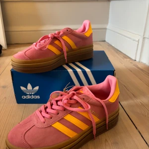 Rosa och gula sneakers från Adidas - Snygga rosa sneakers från Adidas med gula detaljer och en brun sula. Skorna har snörning och en klassisk Adidas-design med de tre ränderna på sidan. Perfekta för en färgglad stil. Skorna är aldrig använda.