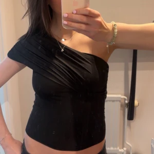 Svart oneshoulder topp - Snygg svart oneshoulder topp med en tight passform. Perfekt för en kväll ute eller en speciell tillställning. Toppen har en elegant och stilren design som framhäver axlarna.