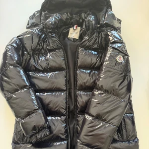 Moncler Maya (NFC)  - Snygg svart glansig dunjacka från Moncler med huva och dragkedja. Jackan har en quiltad design och är perfekt för kyligare väder. Den har en klassisk Moncler-logga på ärmen och en praktisk innerficka med röd dragkedja. NFC Funkar dessutom! 