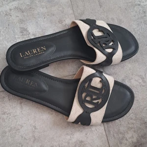 Beige och svarta sandaler från Ralph Lauren - Snygga sandaler från Ralph Lauren med en beige rem och svart logotypdetalj. Perfekta för en stilren look under varma dagar. Sandalerna har en öppen tå och en bekväm sula.