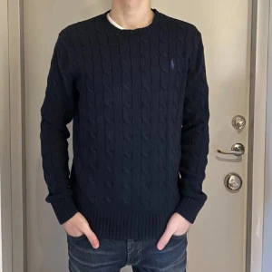 Mörkblå stickad tröja från Ralph Lauren - Passar en vanlig S. Säljer en stilren mörkblå stickad tröja från Ralph Lauren med kabelstickat mönster. Tröjan har långa ärmar och en liten broderad logotyp på bröstet. Perfekt för kyligare dagar!