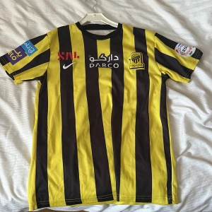 Randig fotbollströja från Nike - Säljer en gul och svart randig fotbollströja från Nike med korta ärmar. Tröjan har Ittihad Club-logga på bröstet och flera sponsorloggor. Perfekt för fotbollsfans som vill visa sitt stöd för laget.