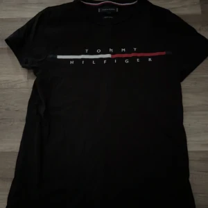Svart t-shirt från Tommy Hilfiger - Snygg svart t-shirt från Tommy Hilfiger med deras ikoniska logga i vitt och rött på bröstet. Perfekt för en stilren look. T-shirten har korta ärmar och en klassisk passform.