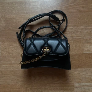 Svart axelväska + förvaringssäck  - Snygg svart axelväska från Charles & Keith med quiltad design och gulddetaljer. Väskan har en justerbar axelrem och ett coolt spänne framtill. Båda remmar går att ta av och justera. Perfekt för att ge din outfit en stilfull touch.