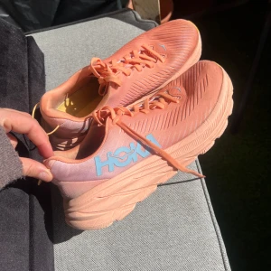 Korallfärgade löparskor från Hoka - Säljer ett par snygga korallfärgade löparskor från Hoka med ljusblå logga på sidan. Skorna har snörning och en bekväm, vadderad sula för extra komfort under löpningen. Perfekta för både träning och vardagliga aktiviteter. Är använda men i mycket bra skick 