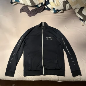 Mörk blå jacka från Hugo Boss - Snygg svart jacka från Hugo Boss med dragkedja och vita detaljer. Jackan har långa ärmar med ett stilrent mönster längs sidorna och ribbade muddar. Perfekt för en casual look.