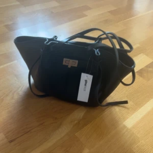 Svart axelväska med metallspänne - Snygg svart axelväska med ett stilrent metallspänne framtill. Väskan har en modern design med långa axelremmar och en praktisk storlek för vardagsbruk. Perfekt för att bära med sig det nödvändigaste med stil.