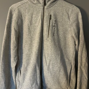 Grå hoodie från Sail Racing  - Säljer en grå hoodie från Sail Racing med dragkedja framtill. Hoodien en diskret logga på bröstet. Samt en liten fläck längst ner. Lite sliten längst ut på armarna. 100kr