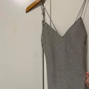 Glittrig silverklänning med smala axelband -  kort glittrig silverklänning med smala axelband och v-ringning. Perfekt för en festlig kväll. Klänningen har en elegant och figurnära passform.Kommer över Knät.