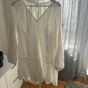 Vit klänning från H&M - Säljer en luftig vit klänning från H&M med v-ringning och långa ärmar. Klänningen har en volangdetalj vid nederkanten och knytband vid halsen. Perfekt för varma dagar!