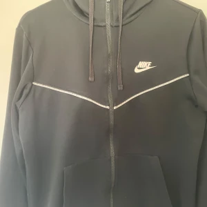 Svart hoodie från Nike - Snygg svart hoodie från Nike med dragkedja och vit logga på bröstet. Den har en klassisk design med justerbar huva och fickor framtill. Perfekt för en avslappnad stil.
