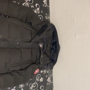 Canada Goose Jacka Svart - Säljer en svart dunjacka från Canada Goose med huva och dragkedja. Jackan har en klassisk design med varumärkets logga på ärmen. Perfekt för kalla vinterdagar.