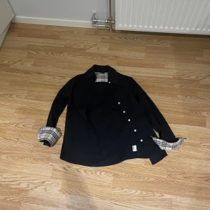 Svart overshirt med rutigt foder - Snygg svart overshirt med ett stilrent rutigt foder i beige och brunt. Jackan har långa ärmar och knappar framtill. Perfekt för lager-på-lager-stilen.