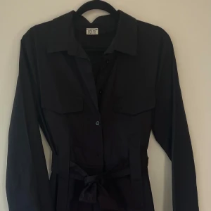 Svart lång jumpsuit från BOMÉ - Snygg svart lång jumpsuit från Bohem med långa ärmar och knappar framtill. Den har en knytning i midjan för en smickrande passform och två bröstfickor som ger en cool touch. Perfekt för en stilren look.