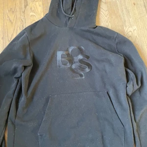Svart hoodie från BOSS - Snygg svart hoodie från BOSS med en stor logotyp på bröstet. Tröjan har en klassisk känguruficka och en bekväm huva med dragsko. Perfekt för en avslappnad stil. Den har inga slitage på sig.