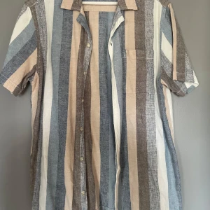 Randig skjorta från Kappahl - Säljer en snygg randig skjorta från Kappahl i storlek M. Skjortan har korta ärmar och ett mönster i jordnära färger som beige, blå och grå. Perfekt för en avslappnad stil.