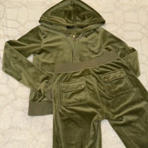 Olivgrön velour hoodie och byxor från Juicy Couture - Säljer en olivgrön velour hoodie och matchande byxor från Juicy Couture. Hoodien har dragkedja och långa ärmar, medan byxorna har praktiska fickor och resår i midjan. Perfekt för en avslappnad och stilren look.