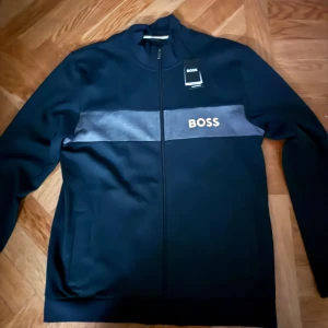 Svart jacka från Boss - Snygg svart jacka från Boss med dragkedja och en grå rand över bröstet där märket är tryckt. Perfekt för en stilren look.