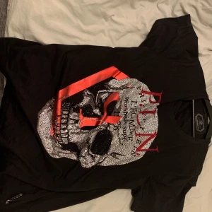 Svart t-shirt med dödskallemotiv från Philipp Plein - Snygg svart t-shirt från Philipp Plein med ett stort dödskallemotiv i silver och rött på framsidan. T-shirten har korta ärmar och en normal passform. Perfekt för dig som vill sticka ut med en cool och edgy stil.