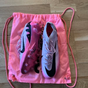 Vita fotbollsskor från Nike - Säljer ett par vita Nike fotbollsskor med svarta detaljer och rosa sulor. Skorna har snörning och är designade för optimal prestanda på planen. Perfekta för fotbollsspelare som vill ha stil och funktionalitet.
