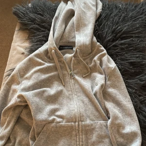 Grå huvtröja från Juicy Couture - Säljer en stilren grå huvtröja från Juicy Couture. Tröjan har dragkedja och en broderad logga på bröstet. Den är tillverkad i en mjuk bomullsblandning och har en justerbar huva med snören. 