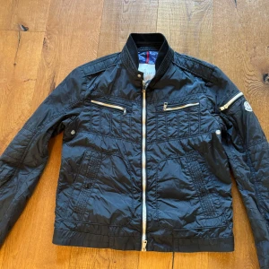 Blå jacka från Moncler - Snygg blå jacka från Moncler med dragkedja och flera fickor. Jackan har en quiltad design och ribbad krage. Perfekt för en stilren look. Storlek 3 motsvarar M hör av er för frågor eller funderingar, väldigt bra skick!