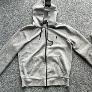  Polo Ralph Lauren Zip Hoodie - Säljer en stilren grå hoodie från Polo Ralph Lauren zip hoodie  med dragkedja och justerbar huva. Tröjan har det klassiska Polo-logotypen broderad på bröstet. Perfekt för en avslappnad stil. Skick 9/10 Inga defekter 