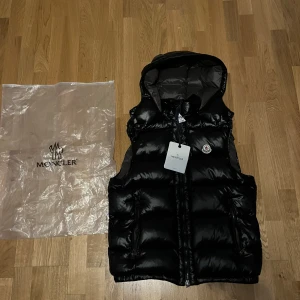 Moncler Väst - Säljer en snygg svart dunväst från Moncler med dragkedja och huva. Storleken är M men är liten i storleken så den passar mer som en S skulle jag säga, men kan även passa M