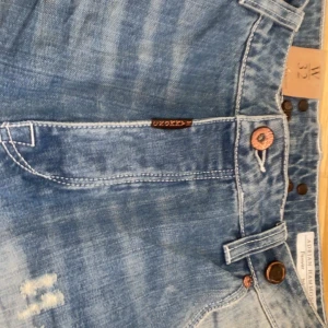 Blå jeans från Adrian Hammond - Snygga blå jeans från Adrian Hammond med slitna detaljer och knappgylf. Perfekta för en avslappnad stil. De har en klassisk femficksdesign och är tillverkade i ett bekvämt denimtyg. Aldrig använda allt är kvar på byxorna o dem säljs inte längre 