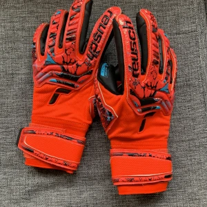 Röda fotbollshandskar från Reusch - Snygga röda fotbollshandskar från Reusch med svarta och blå detaljer. Handskarna har en greppvänlig yta och justerbara kardborreband vid handleden för perfekt passform. Perfekta för målvakter som vill ha stil och funktionalitet på planen.