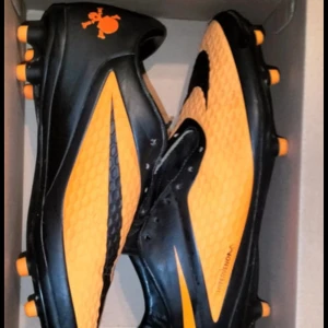 Nike hypervenom - Snygga fotbollsskor från Nike i svart och orange med en cool dödskalle-detalj. Skorna har en rund tå och är designade för att ge bra grepp på planen med sina orange dobbar. Perfekta för fotbollsträning eller match.