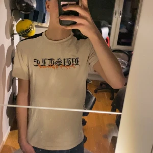 Beige t-shirt från Supply & Demand - Snygg beige t-shirt från Supply & Demand med texten 'HUSTLE' i svart och orange på framsidan. T-shirten har svarta detaljer längs axlarna och är kortärmad. Perfekt för en avslappnad stil. Väldigt sparsamt använd. Skulle säga att den passar bäst på 170-175!